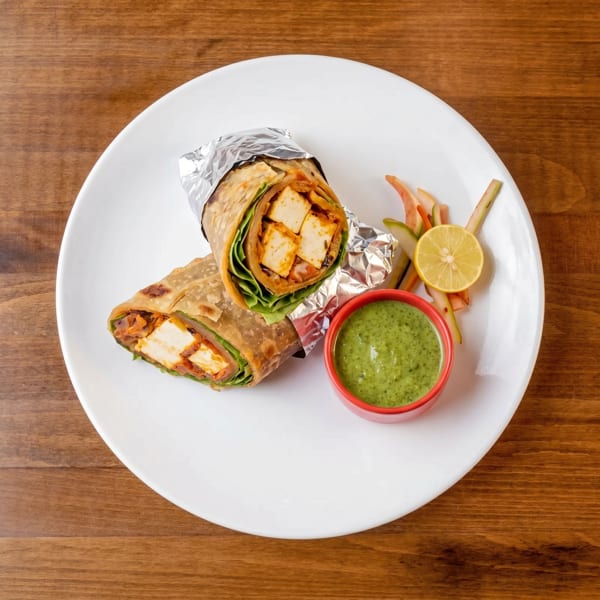 Two paneer wraps, one partially unwrapped, with green chutney and lemon on a white plate.