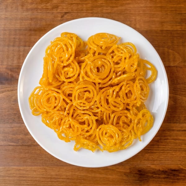 A white plate filled with golden-orange jalebi, a spiral-shaped Indian sweet, on a wooden table.