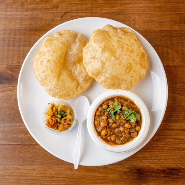 Two golden-brown puri breads, a bowl of chana masala, and a small container of pickle on a white plate.
