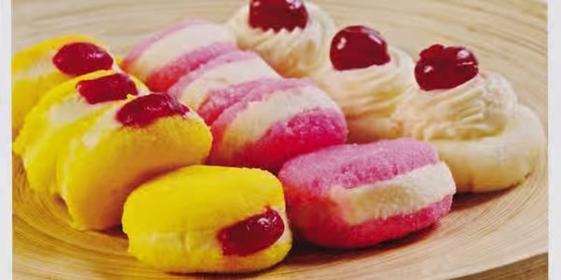 A wooden plate filled with colorful Indian sweets, including pink and yellow sandesh with cherry toppings.