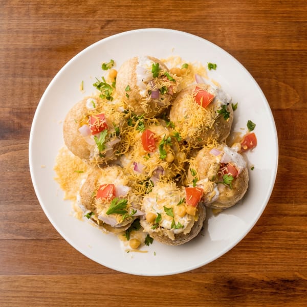 A white plate holds several pani puri, topped with yogurt, chopped tomatoes, onions, and cilantro.