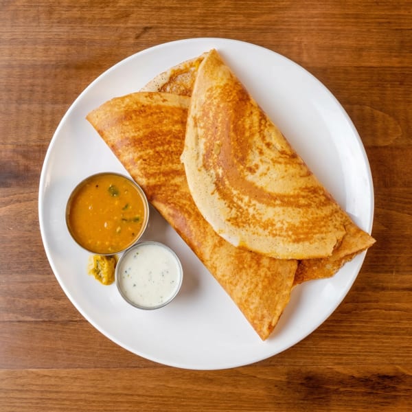 Two golden-brown dosas, folded, served with sambar and chutney in small metal bowls on a white plate.