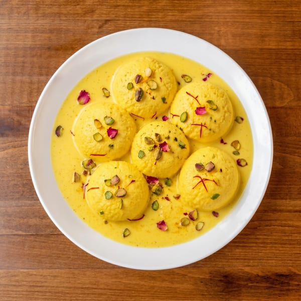 Rasmalai, an Indian dessert, with pistachios, saffron, and rose petals in a creamy yellow sauce.